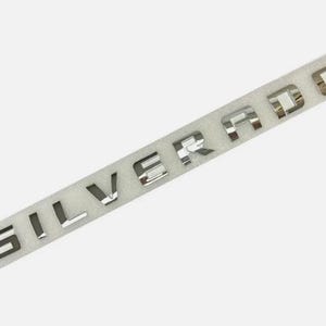 May include: Silver chrome letters spelling "SILVERADO" on a white foam strip. The letters are reflective and appear to be made of metal. The letters are evenly spaced and are in a sans-serif font. The background is a plain white.
