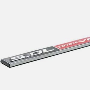 Puede incluir: Un emblema rectangular plateado con el texto "5.0L" en blanco y "COYOTE V8" en rojo. El emblema tiene un acabado metálico y probablemente sea para uso automotriz.