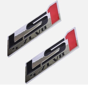Puede incluir: Dos emblemas LS1 cromados con letras negras y un acento rojo. Los emblemas rectangulares presentan el texto "LS1 5.7L V8". El diseño es una representación estilizada del tipo de motor.