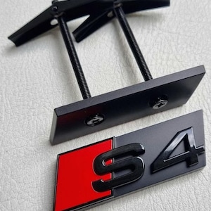 May include: Black metal car emblem with the letters "S4" in black on a grey background, with a red square on the left. Includes a mounting bracket with two threaded rods and a black metal plate.
