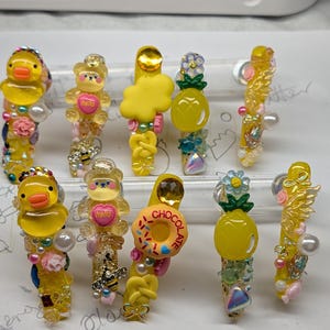 May include: Ten yellow hair clips decorated with resin charms including ducks, teddy bears with the word hello, pineapples, donuts with the word chocolate, clouds, and pretzels. Each clip features various beads, pearls, and small floral accents.