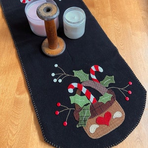 May include: Black felt table runner with a stitched design of a brown basket filled with candy canes and holly. The runner has red berries and white dots stitched on it. Two candles are on the runner, one pink and one white.