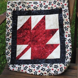 May include: A quilted tote bag with a red and white star pattern, black border, and cream-coloured handles. A white hat with a bow rests on a wooden chair. The bag features a repeating pattern of black figures and red maple leaves.