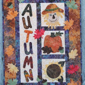 Pode incluir: Uma tapeçaria de parede acolchoada com tema de outono. A palavra "AUTUMN" é exibida verticalmente. A colcha apresenta um espantalho, uma abóbora e um girassol, rodeados por folhas coloridas. A paleta de cores inclui laranja, castanho e roxo.