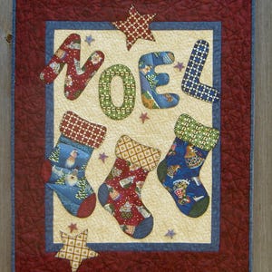 Peut inclure: Une tenture murale de Noël matelassée avec le mot "NOEL" et trois chaussettes. Les lettres et les chaussettes sont en tissu à motifs rouge, vert, bleu et or. Le fond est crème, avec une bordure rouge et des étoiles.