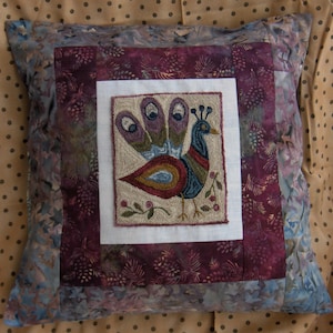 Könnte beinhalten: Quadratisches Zierkissen mit einem farbenfrohen, gestickten Pfauendesign. Das Kissen hat ein zentrales Quadrat mit dem Pfau, umgeben von einem weißen Rand, einem dunkelburgunderfarbenen Quadrat und einem gemusterten blauen Rand.