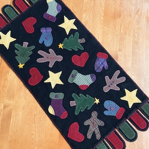 May include: A black felt table runner with a Christmas theme. It features red, green, and yellow felt appliques of hearts, stars, mittens, trees, stockings, gingerbread men, and reindeer. The runner has a fringe of red and green felt.