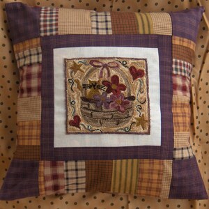 May include: Square decorative pillow with a patchwork design. The center features a stitched image of a basket with flowers and a cat. The pillow has a purple border and a variety of plaid and patterned fabric squares.