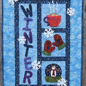 May include: A quilted wall hanging with a winter theme. The word "WINTER" is spelled out vertically in purple letters. The design includes a red mug of hot cocoa, mittens, snowflakes, and a snow globe. The background is light blue.