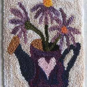 Könnte beinhalten: Handgefertigte Blumen-Wandbehänge mit einer lila Gießkanne mit einem rosa Herzen und lila Blumen. Der Hintergrund ist beige mit einem karierten Rand in Lila und Marineblau. Die Blumen haben orange und gelbe Zentren.