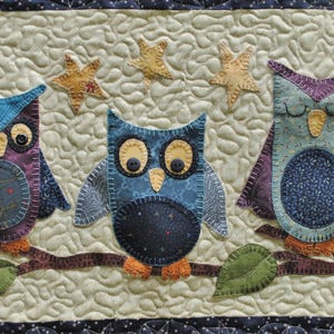 May include: A quilted wall hanging featuring three whimsical owl figures perched on a branch. The owls are in shades of blue, purple, and teal, with button eyes and decorative stitching. Beige stars are scattered above them against a quilted background.