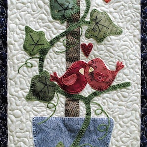 May include: A quilted wall hanging featuring a tree with leaves, two red birds, a butterfly, and a blue pot. The background is white with a quilted pattern. The border is dark blue with white stars.