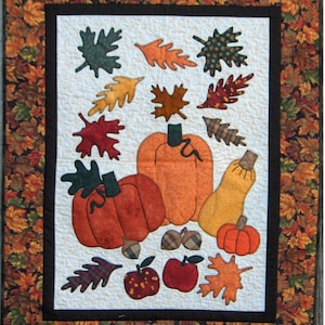 May include: A quilted wall hanging with autumnal appliqués. The design features pumpkins, gourds, apples, acorns, and leaves in orange, red, green, and brown. The central panel is framed in black and surrounded by a border of leaf-patterned fabric.
