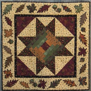 Könnte beinhalten: Eine handgefertigte Steppdecke mit einem zentralen Sterndesign in Grün-, Orange- und Burgundertönen. Die Steppdecke hat einen Rand aus applizierten Blättern und Eicheln auf cremefarbenem Hintergrund, wodurch ein Herbstthema entsteht.