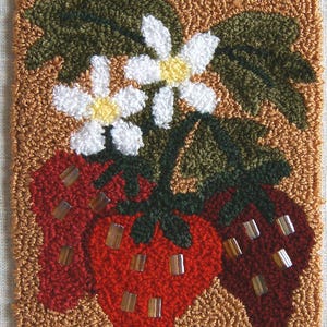 Könnte beinhalten: Ein handgenähter Teppich mit Erdbeer-Design. Der Teppich zeigt drei rote Erdbeeren mit transparenten Perlenakzenten, weißen Blüten und grünen Blättern. Der Rand hat ein kariertes Muster in Gelb und Marineblau.