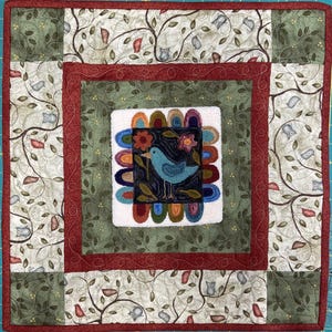 May include: A square, handmade quilt with a central design of a blue bird on a dark background, surrounded by colorful arches and flowers. The quilt features a green and cream owl and leaf pattern, framed by a red border. The overall design is a patchwork style.