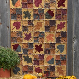 Może przedstawiać: Pikowana narzuta w stylu patchwork z motywem jesiennym, z kwadratami z różnych tkanin w odcieniach złota, brązu i bordo. Narzuta jest ozdobiona aplikacjami liści i żołędzi w jesiennych kolorach. Narzuta jest prezentowana na rustykalnym drewnianym tle.