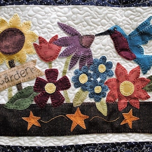 Może przedstawiać: Pikowana scena ogrodowa z różnorodnymi kolorowymi kwiatami z tkaniny, kolibrem i znakiem z napisem "Garden". Kwiaty znajdują się w ciemnobrązowej donicy z pomarańczowymi gwiazdkami. Tło jest białe z pikowanym wzorem.