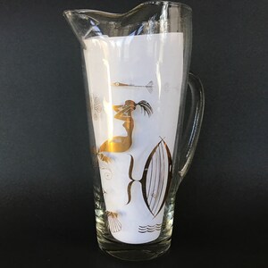 May include: A clear glass pitcher with a gold design of a woman and fish. The pitcher has a white background and a gold rim.