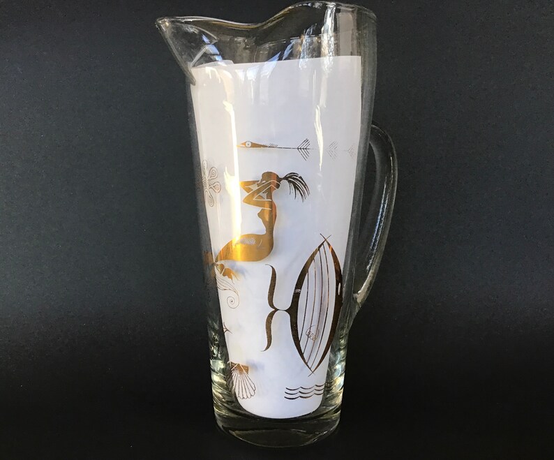 May include: A clear glass pitcher with a gold design of a woman and fish. The pitcher has a white background and a gold rim.
