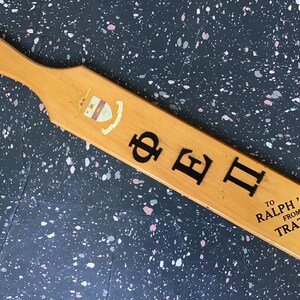 May include: Wooden paddle with a handle and a hole. The paddle is light brown with black lettering and a crest. Text includes "ΦEI", "To Ralph '69 From Tratt '69".