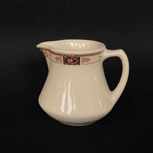 May include: Cream-colored ceramic creamer with a decorative rim featuring a repeating pattern in brown and black. The creamer has a curved body, a handle, and a spout for easy pouring. A classic piece for serving cream or milk.