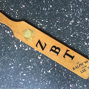 May include: Wooden paddle with a handle and a hole. The paddle has the letters "NBT" in black, along with a crest and the inscription "TO RALPH '69 FROM LEE '69".