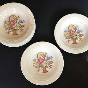 May include: Set of three white ceramic bowls with a floral design. Each bowl features a basket of flowers with a cross-stitch pattern.
