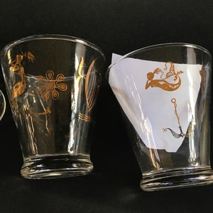 May include: Three clear glass tumblers with gold-colored mermaid and sea-life designs. The tumblers are on a black surface.
