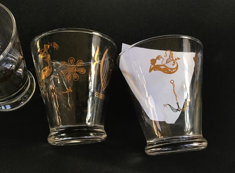 May include: Three clear glass tumblers with gold-colored mermaid and sea-life designs. The tumblers are on a black surface.