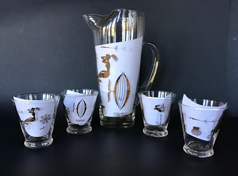 May include: A vintage glass pitcher and four matching glasses with gold and black nautical designs. The pitcher has a handle and a wide mouth. The glasses are short and have a wide base.