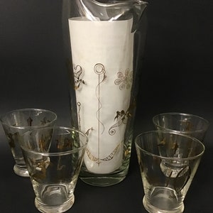 May include: A set of five vintage glass cocktail glasses with gold accents. The glasses feature a stylized design of a bird, a flower, and a spiral. The pitcher is tall and cylindrical with a wide base.