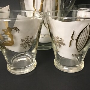 May include: Set of four clear glass tumblers with gold trim and a white background. The glasses feature a mermaid, a starfish, and a sailboat design.