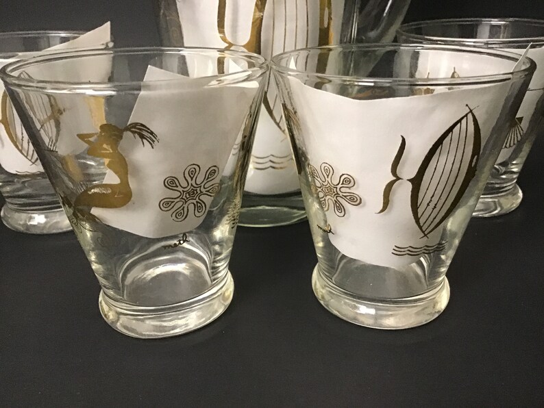 May include: Set of four clear glass tumblers with gold trim and a white background. The glasses feature a mermaid, a starfish, and a sailboat design.
