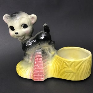 May include: A ceramic planter shaped like a hay bale with a black and white deer figurine standing on top. The deer has large eyes and a pink nose. The planter is yellow with green accents and a pink brick wall behind the deer.