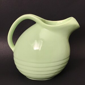 May include: A pale green ceramic pitcher with a rounded body and a slightly flared spout. The pitcher has a smooth, glossy finish and a slightly textured, ribbed design around the bottom.