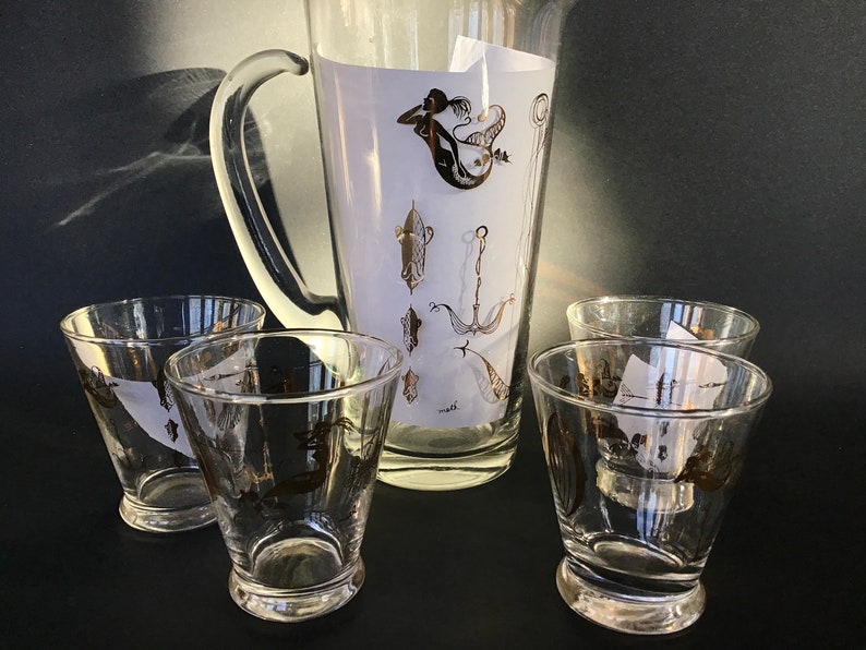 May include: A set of five clear glass drinking glasses with gold nautical designs, including a mermaid, anchor, and fish. The glasses are part of a pitcher set.