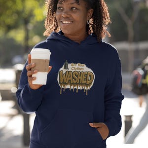 May include: Navy blue hoodie with a front pocket and a graphic that reads "Are Your Clothes Washed" in a brown and white design. The person in the image is holding a white coffee cup with a brown sleeve.