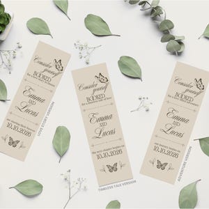 Op de afbeelding: Drie beige bladwijzers met de tekst "Consider yourself BOOKED" en de namen "Emma en Lucas" met een trouwdatum. De bladwijzers zijn versierd met vlinders en bloemmotieven. Groene bladeren en witte bloemen omringen de bladwijzers.
