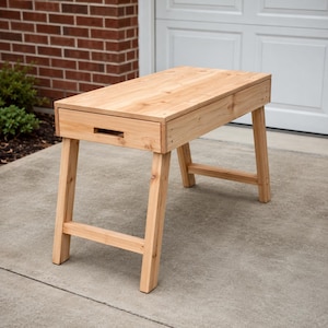 May include: A light-colored wooden desk featuring a rectangular top and a single front-facing drawer with a rectangular cutout handle. The desk is supported by four angled legs connected by horizontal support bars on the sides and back.