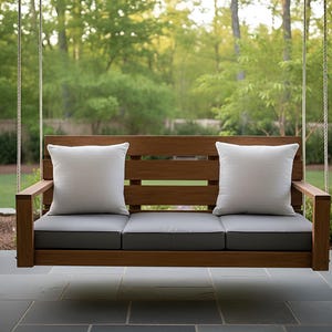 May include: A wooden porch swing with a slatted back and armrests. It has gray cushions and two white square pillows. The swing is suspended by ropes and set against a backdrop of trees.