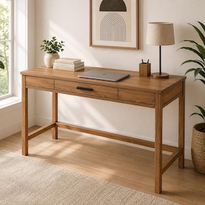 May include: A rectangular wooden desk with a natural brown finish, featuring three front drawers with black horizontal handles. The desk has four straight legs connected by lower support bars and a flat surface holding a laptop and books.