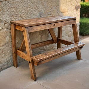 May include: A wooden bar-height table with a natural finish. The table has a rectangular top and a footrest. The legs are angled for stability. The table is made of solid wood and is suitable for outdoor use.