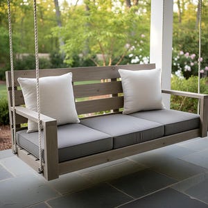 Peut inclure: Un banc-swing en bois avec des coussins gris et des oreillers blancs. Le banc est suspendu par des cordes et possède des accoudoirs. L'arrière-plan présente de la verdure et un patio en pierre, créant un coin salon extérieur relaxant.