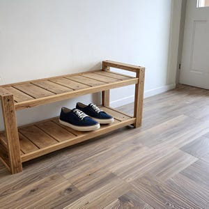 Puede incluir: Un zapatero de madera clara de dos niveles. Dos pares de zapatillas deportivas azules con cordones blancos están en el estante inferior. El zapatero está contra una pared blanca. Las dimensiones del zapatero son en centímetros.