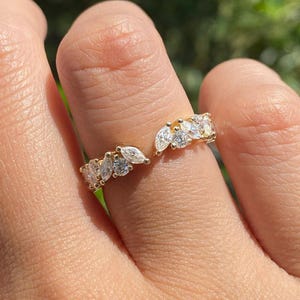 May include: A gold-colored ring with a split design, adorned with multiple clear, sparkling gemstones. The ring features a cluster of marquise-cut and round-cut stones, creating a delicate and elegant appearance. The ring is shown on a finger.