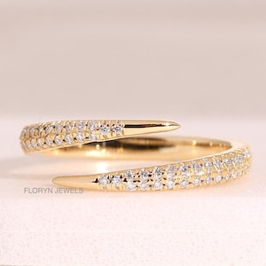 Könnte beinhalten: Ein goldener Bypass-Ring mit zwei spitz zulaufenden Enden, die mit kleinen, klaren Pavé-Steinen besetzt sind. Der Ring hat eine polierte Goldoberfläche und der Text FLORYN JEWELS ist in kleinen schwarzen Buchstaben an der Basis aufgedruckt.