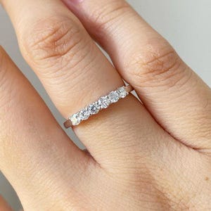 May include: A delicate silver ring featuring a row of seven round, clear gemstones. The ring is set on a slender band and is shown on a person's finger. The gemstones sparkle, reflecting light. The ring is a piece of jewelry.