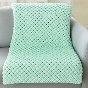 May include: A mint green woven blanket draped over a gray sofa. The blanket has a thick, textured weave pattern. The soft, plush blanket is folded over the sofa's arm and seat, creating a cozy and inviting look. The blanket is a solid color.
