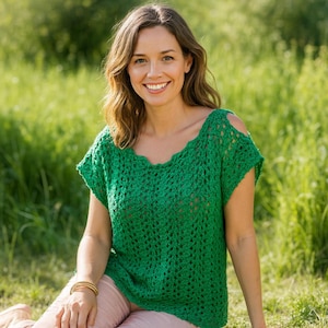May include: A woman wearing a green, open-weave crochet top with short sleeves and a scalloped neckline. The top has cut-out details on the shoulders. She is sitting outside in a grassy area, smiling at the camera.